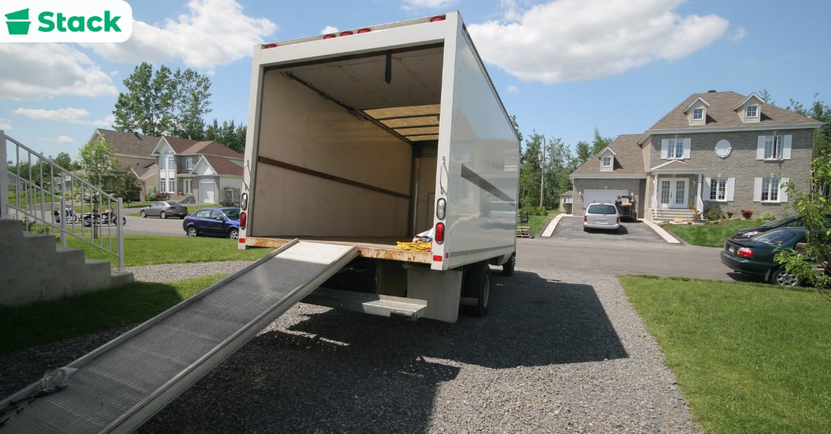 Open moving truck with its loading ramp lowered on a gravel driveway in front of a suburban house